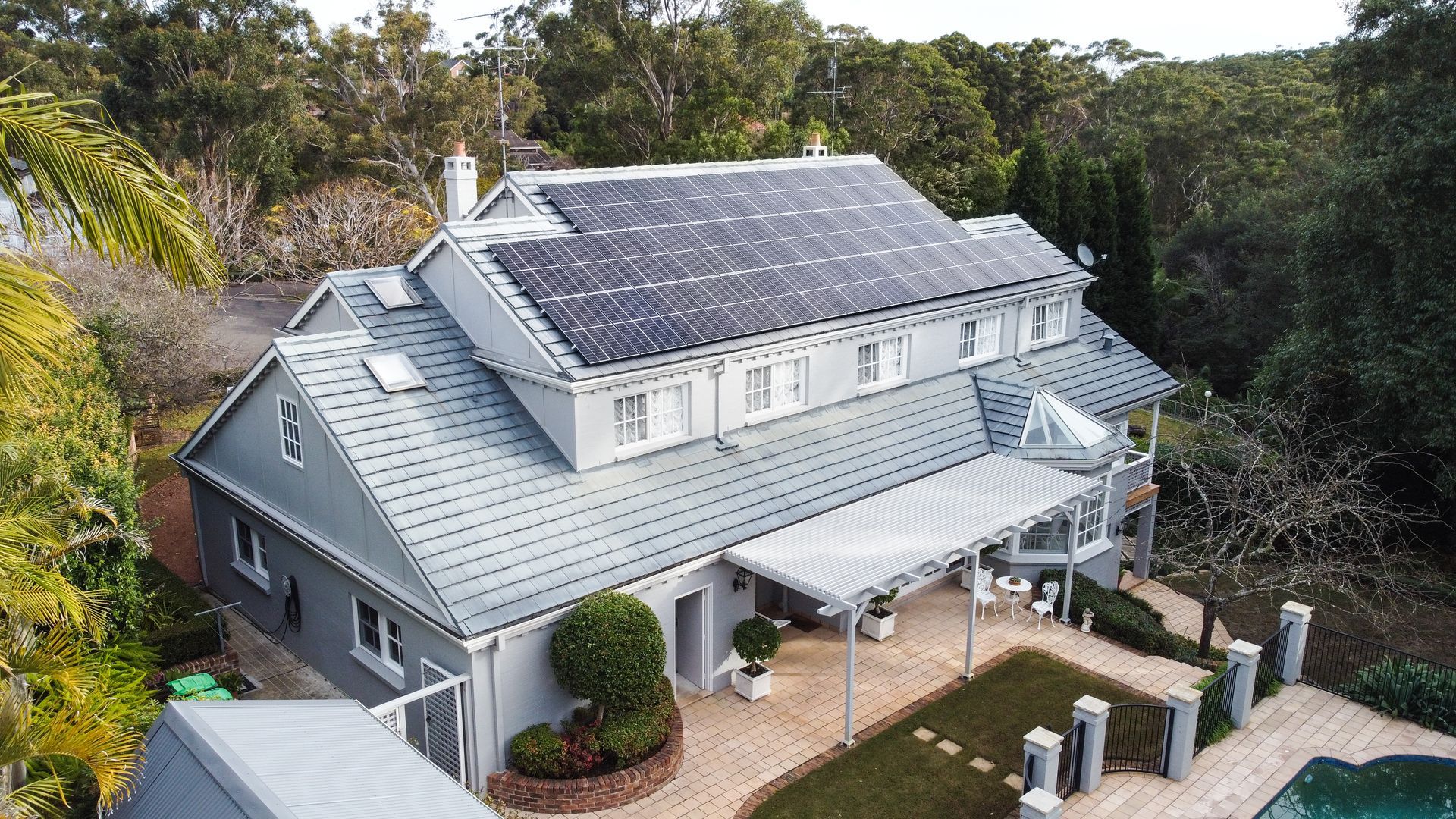 A gray house with solar panels on the roof beside a pool and surrounded by trees.