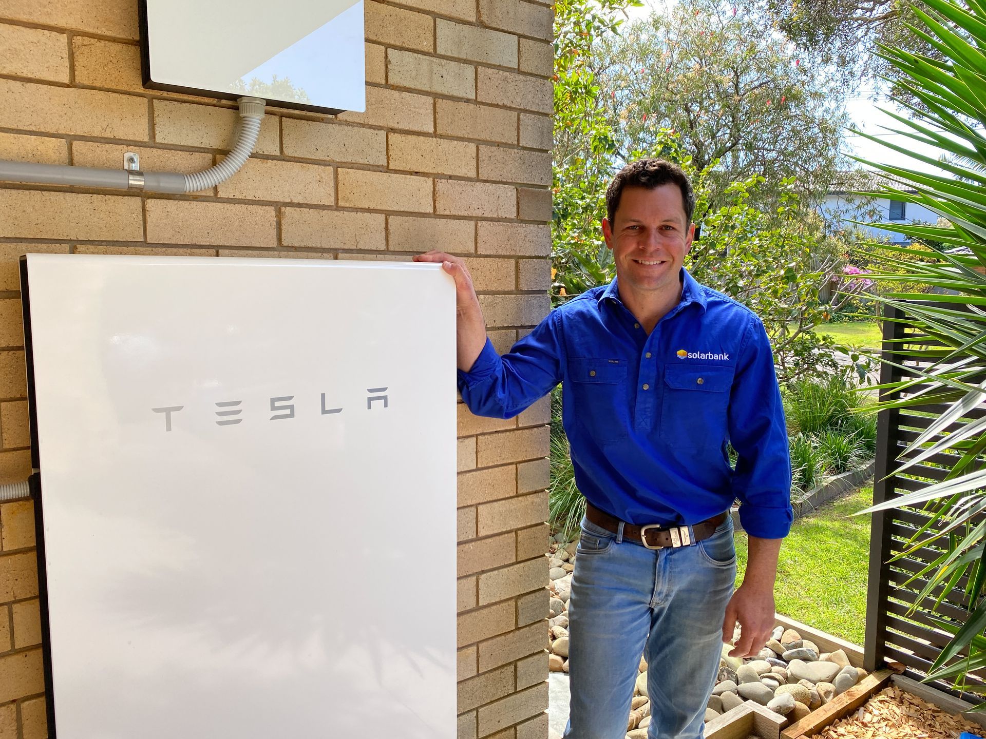 Man in blue shirt stands beside a white Tesla battery unit outdoors by a brick wall.