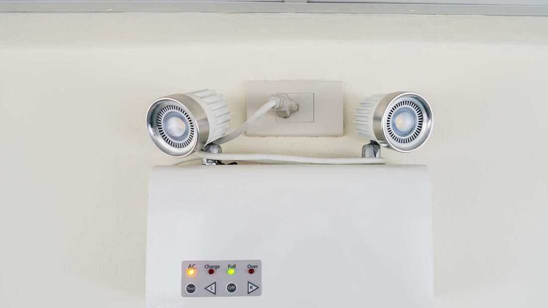 White emergency light fixture with two spotlight heads and status indicator lights on a wall.