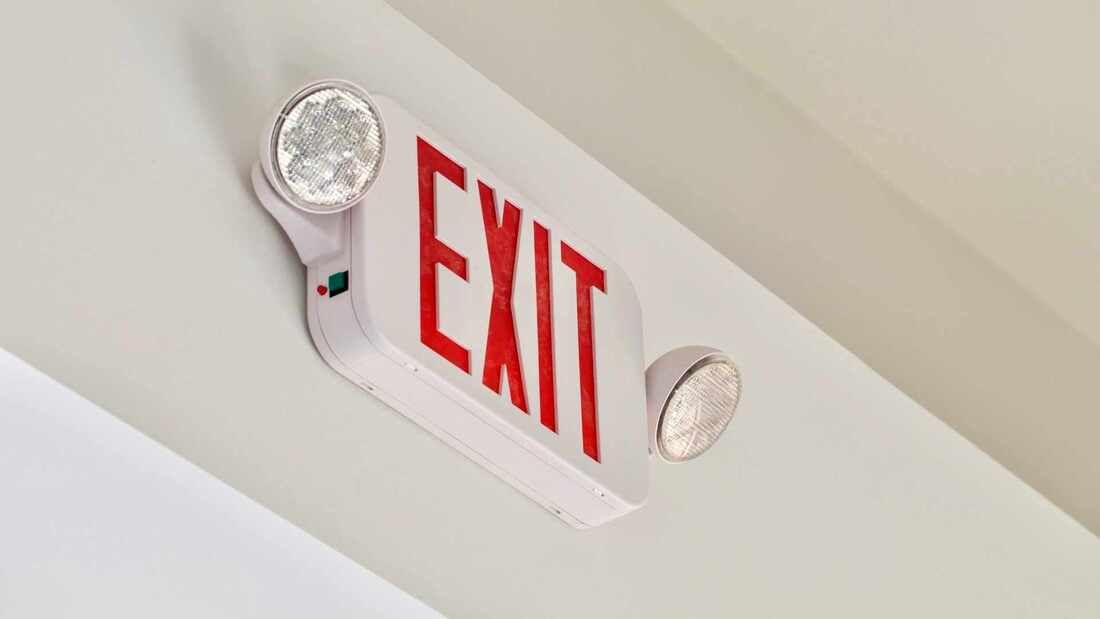 White exit sign with red letters, two lights attached, mounted on a white ceiling.