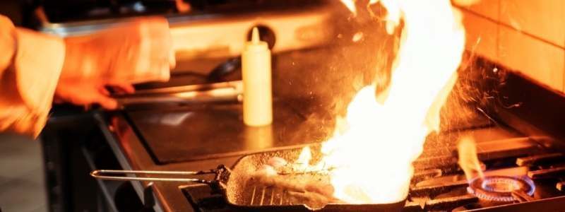 Flames erupt from a pan on a stove in a commercial kitchen; a chef's hand is visible.
