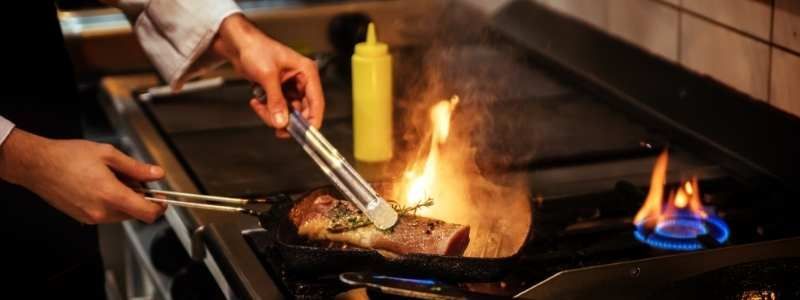 A chef uses tongs to cook meat on a flaming stovetop in a kitchen. A yellow bottle of sauce is nearby.