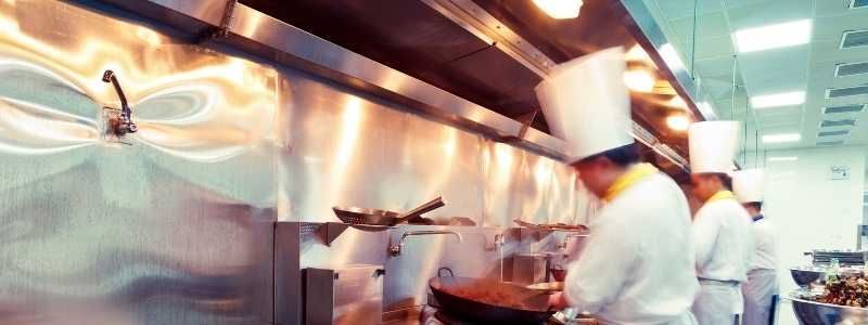 Chefs in white uniforms and hats cooking in a commercial kitchen under a stainless steel hood.
