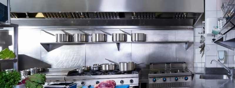 A stainless steel commercial kitchen. Pots sit on a shelf and stovetop. A large exhaust hood hangs above.