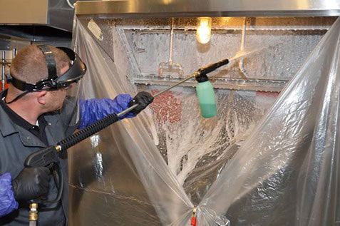 Person cleaning a machine with a spray wand, wearing safety gear, enclosed in plastic sheeting.