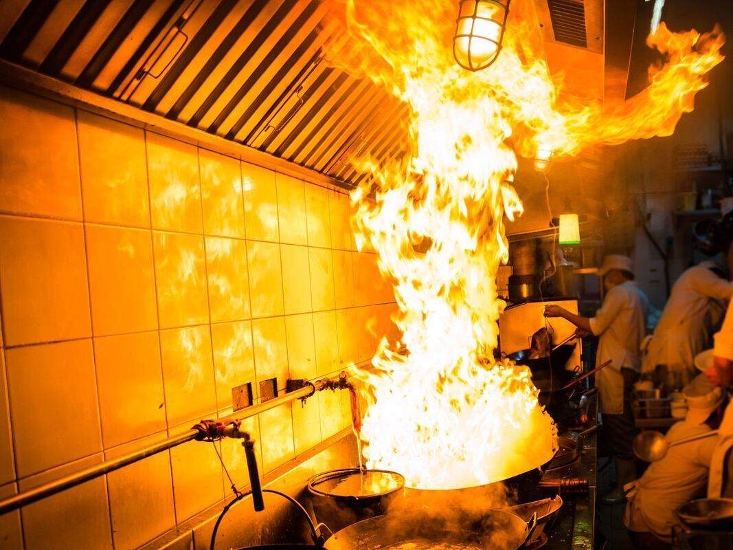 Fire erupts from a wok in a commercial kitchen; flames reach the ceiling.