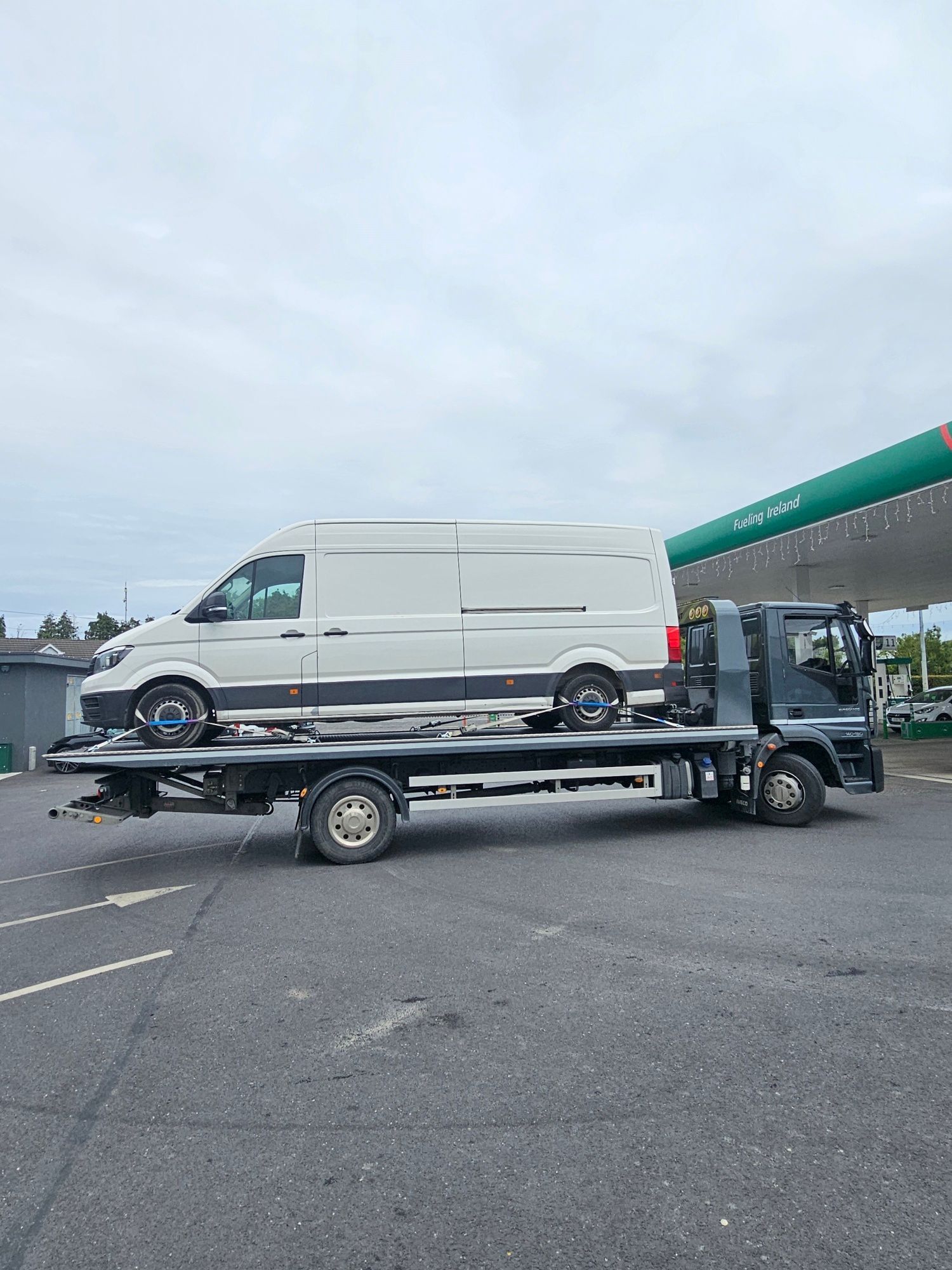 White cargo van on a flatbed tow truck in a parking lot under an overcast sky.