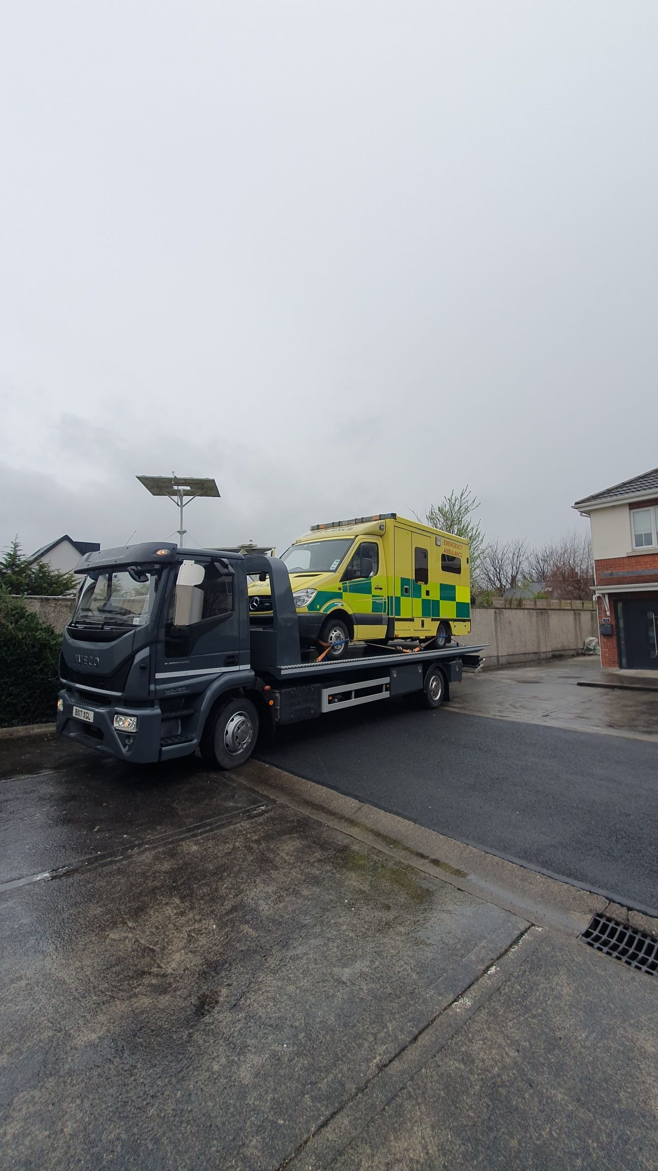 Tow truck carrying a yellow-green ambulance on a wet street under an overcast sky