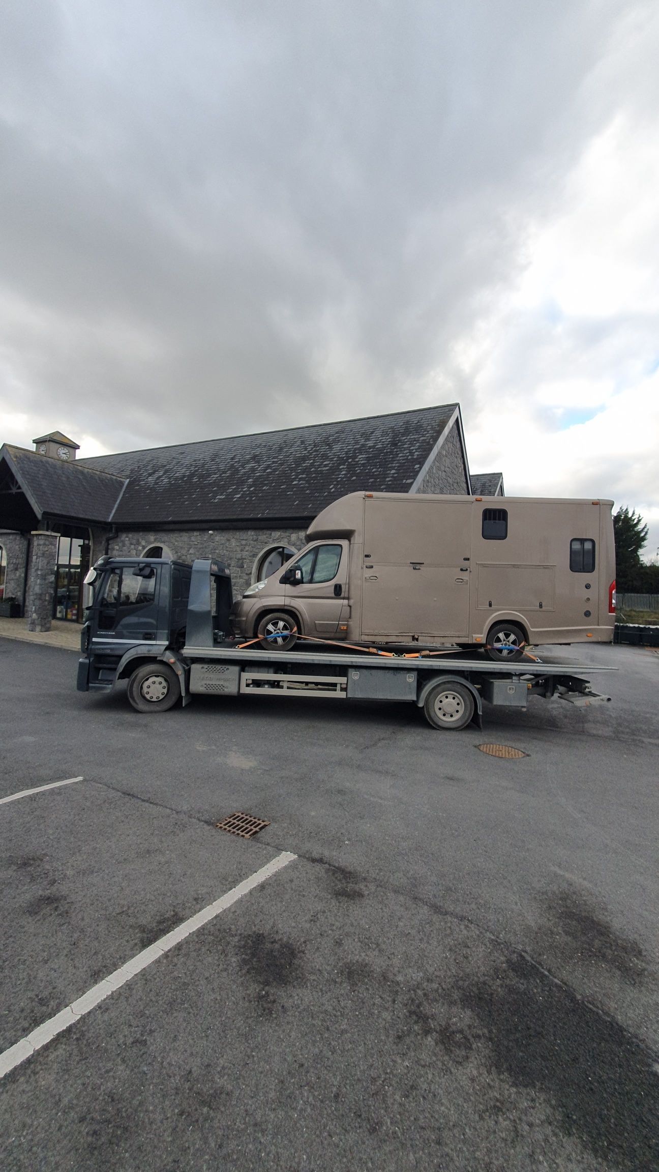 Flatbed truck carrying a tan camper shell in a gravel lot beside a stone building under cloudy skies