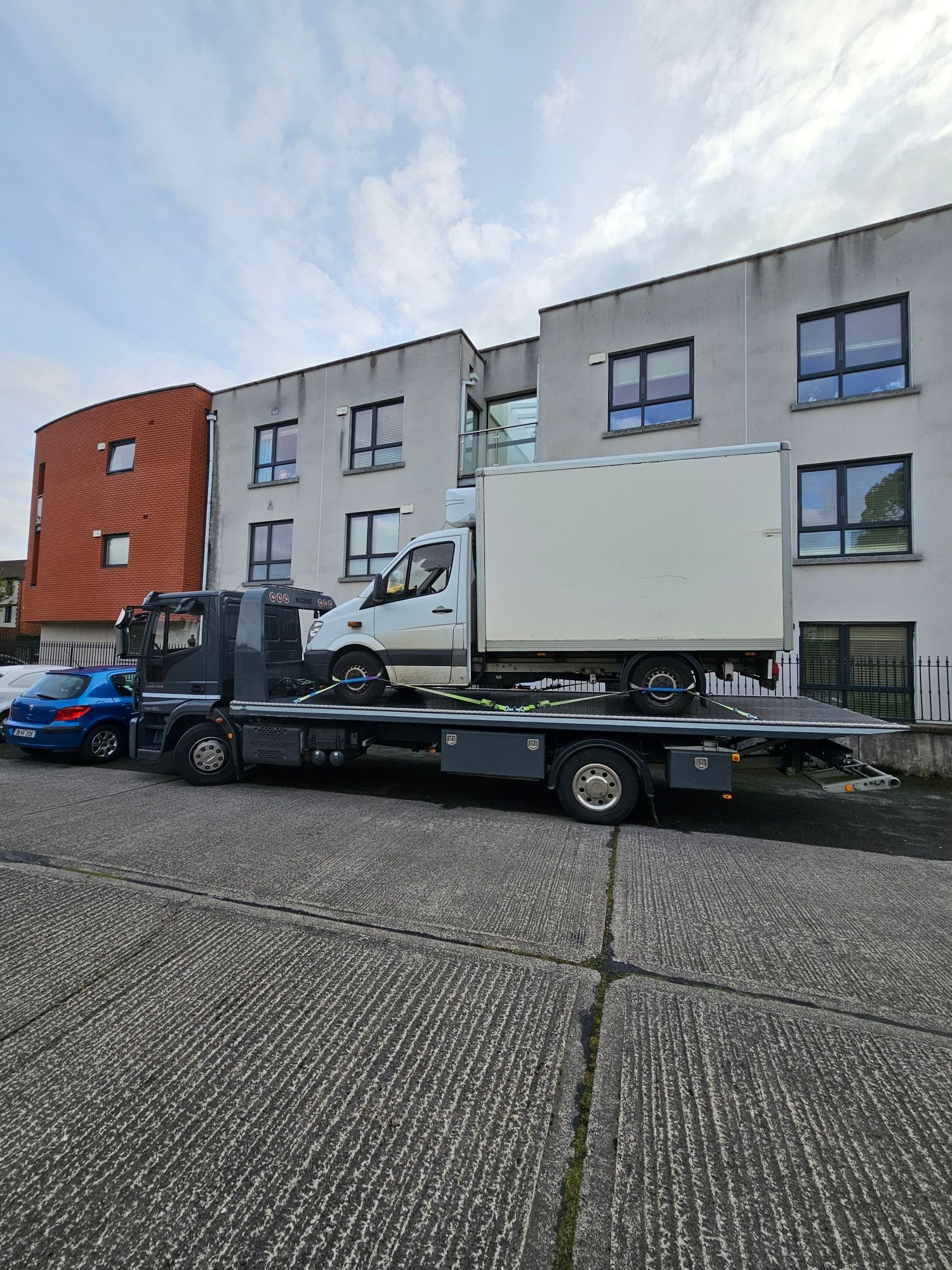 Tow truck carrying a white box truck parked outside a modern apartment building