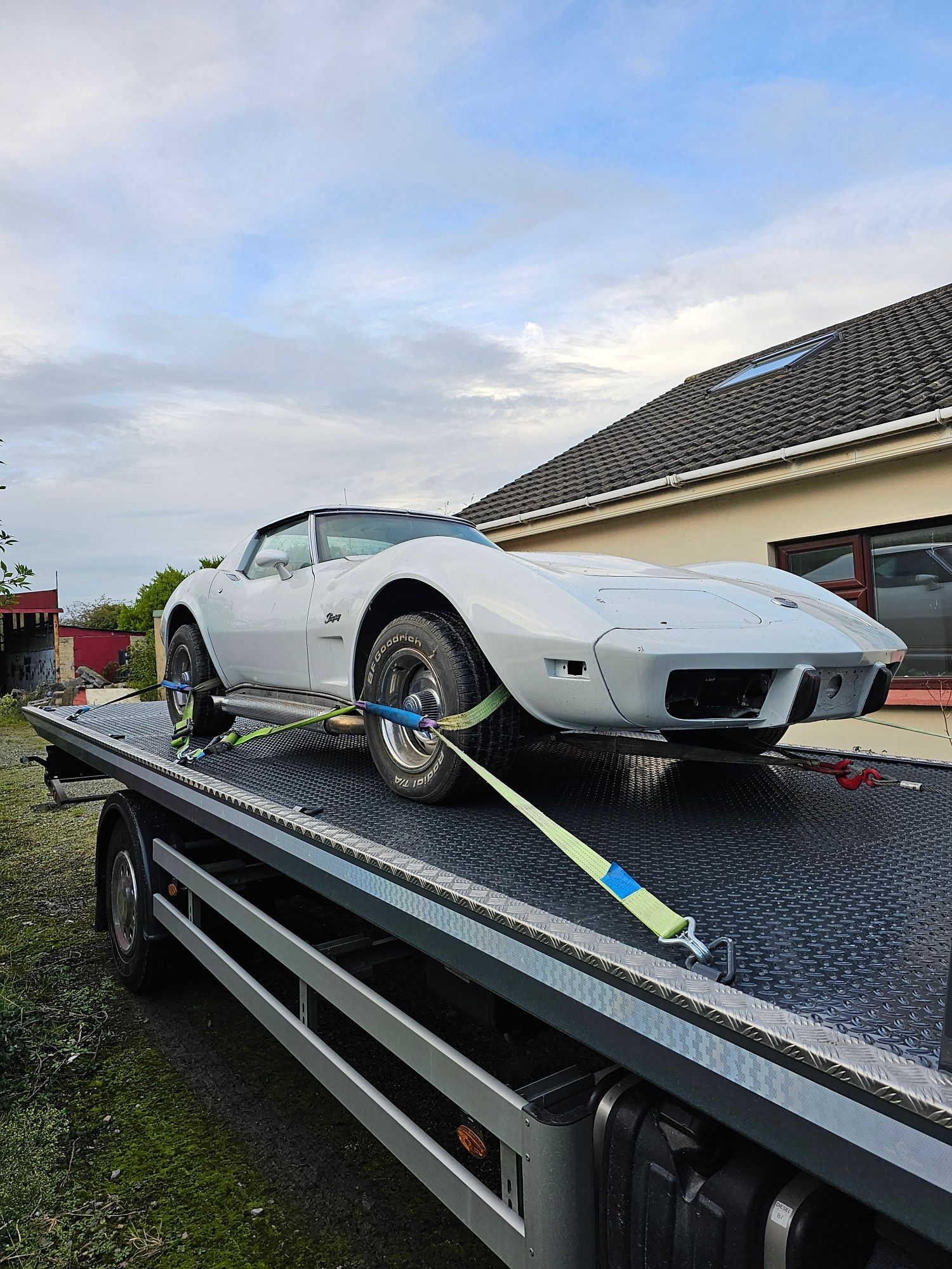 White sports car strapped on a flatbed tow truck in front of a house at dusk