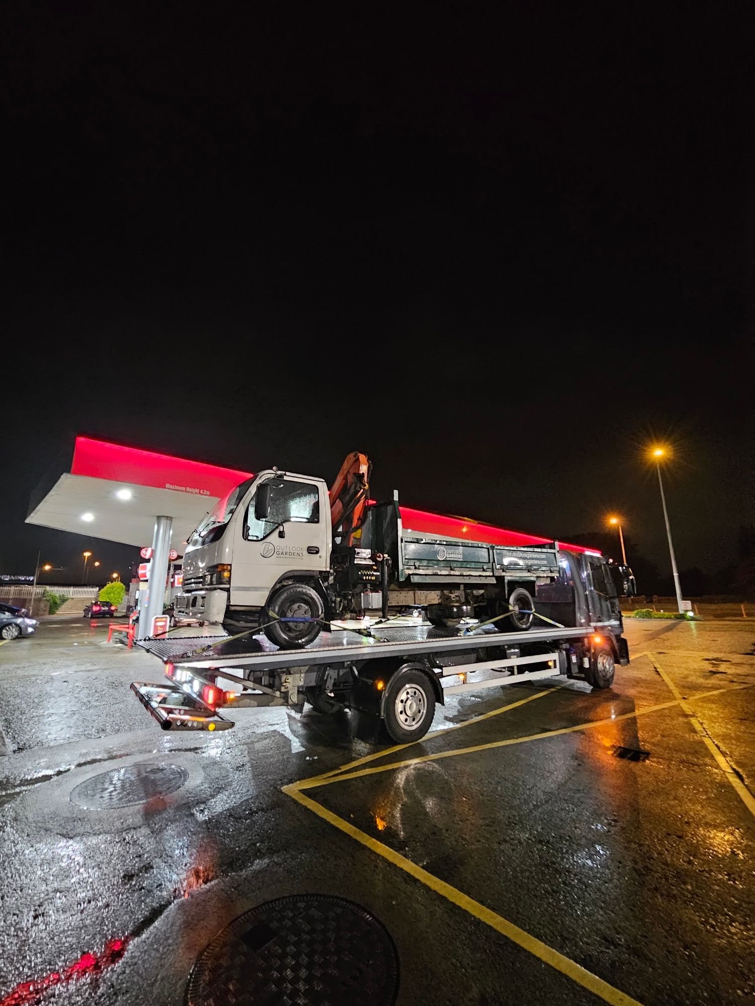 Tow truck loading a white pickup at night in a wet parking lot under bright lights