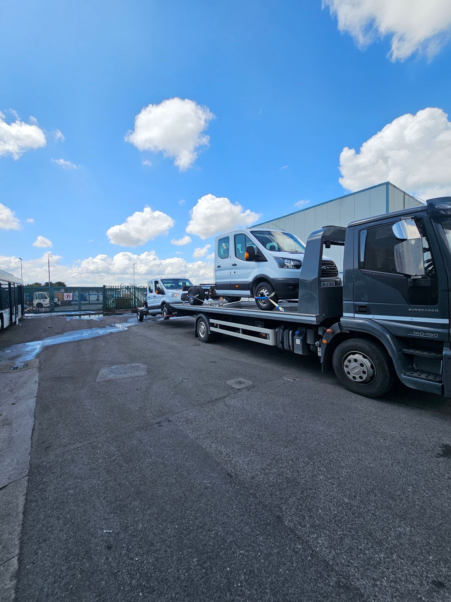 Tow truck carrying a white van on a road under a bright blue sky with clouds
