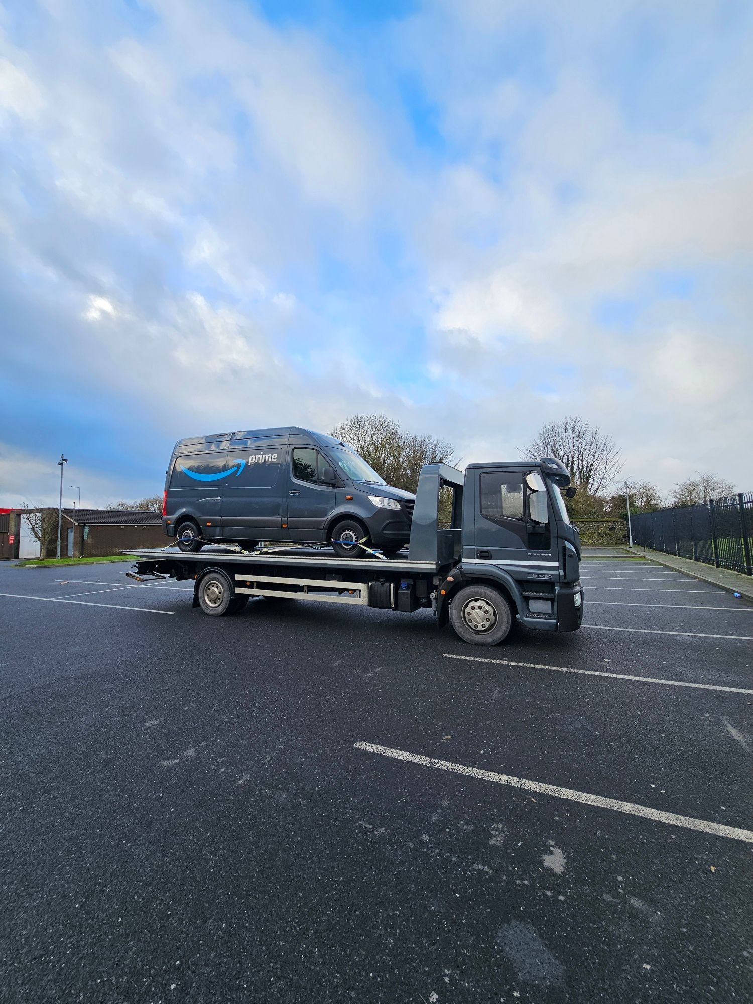 Tow truck carrying a dark van in an empty parking lot under a cloudy sky