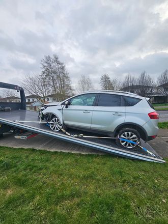 Silver SUV tipped on a tilted trailer ramp in a grassy yard under cloudy skies