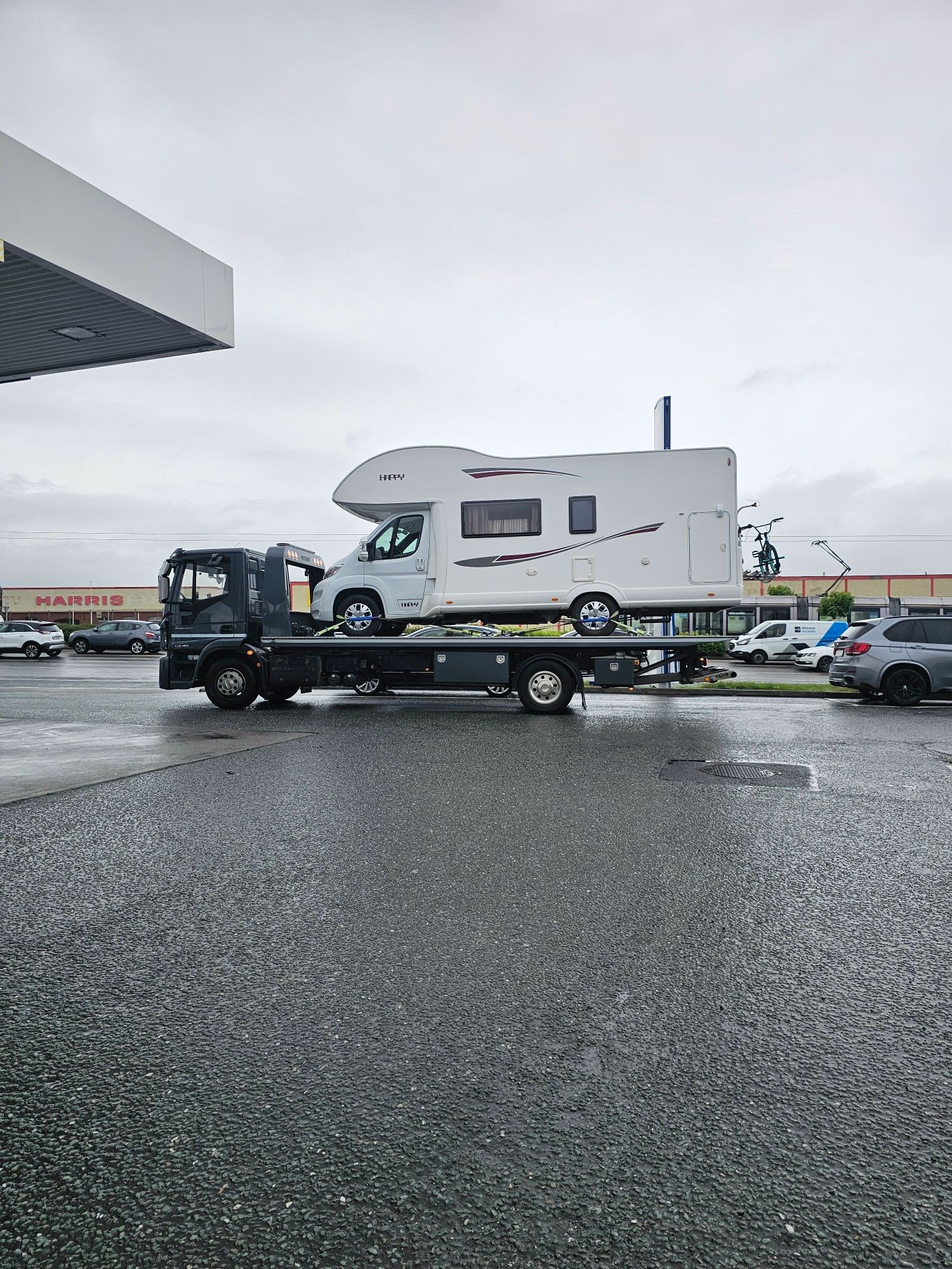 White RV on a flatbed tow truck in a wet parking lot under overcast skies.