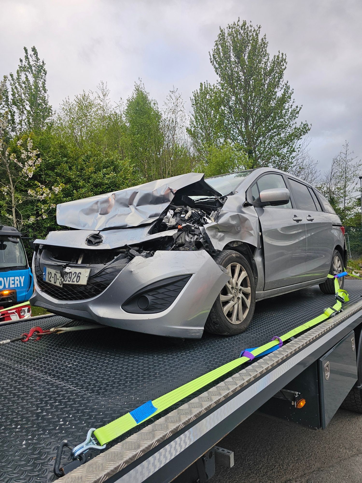 Severely wrecked silver car loaded on a tow truck, front end crushed after a crash.
