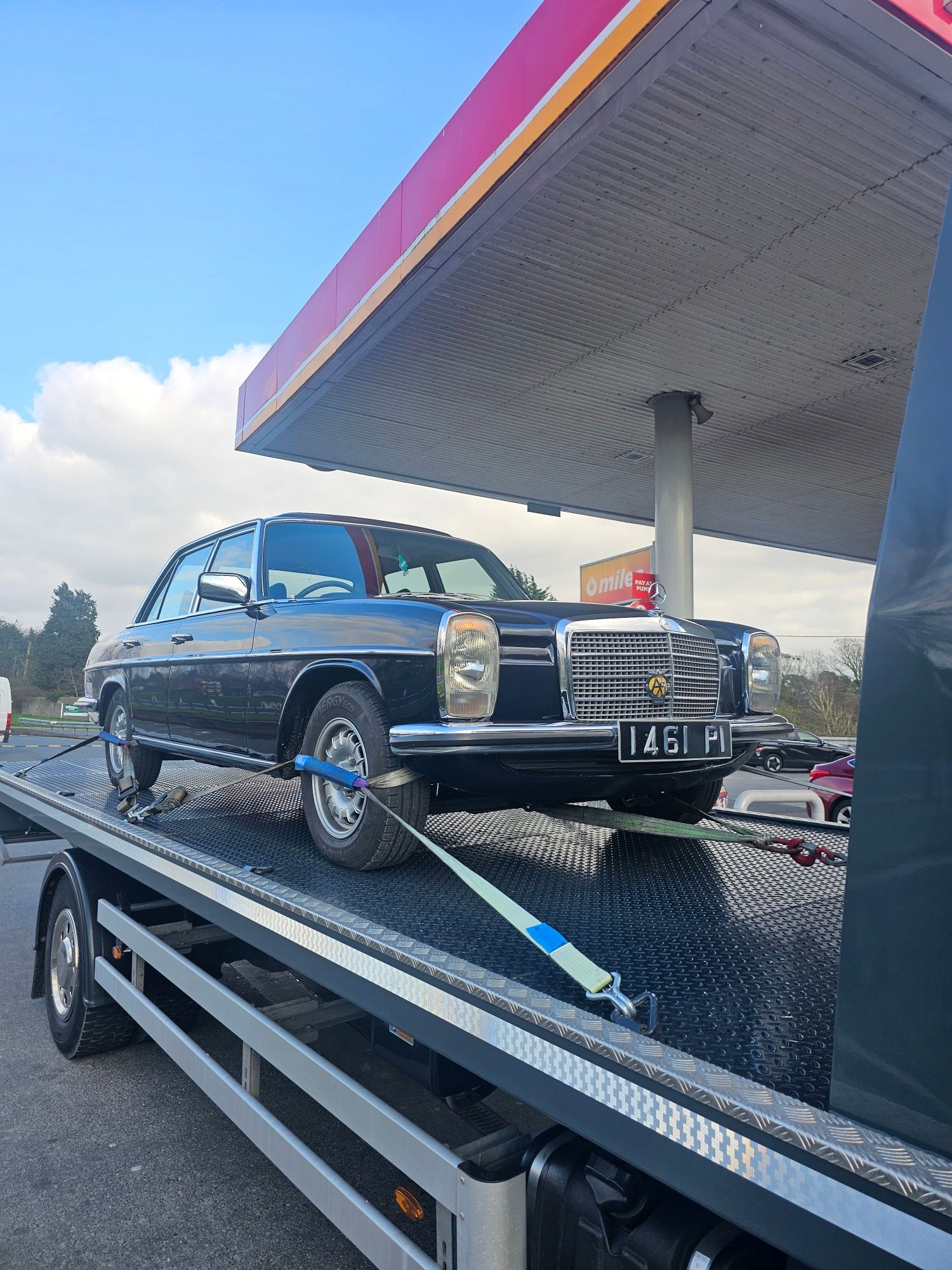 Classic black vintage car on a trailer under a gas station canopy, with blue sky and clouds.