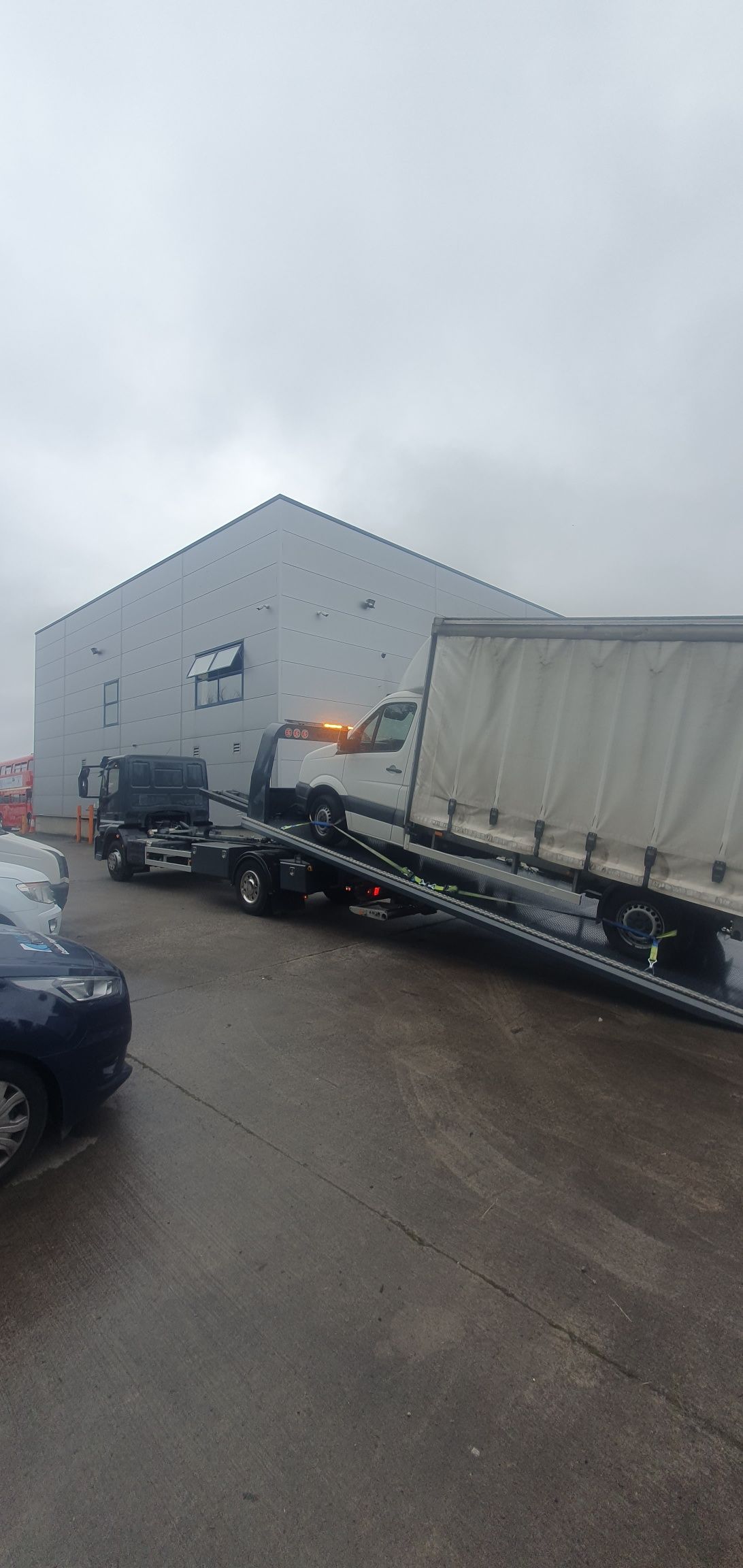 Tow truck loading a white semi-trailer on a wet roadside beside a gray industrial building