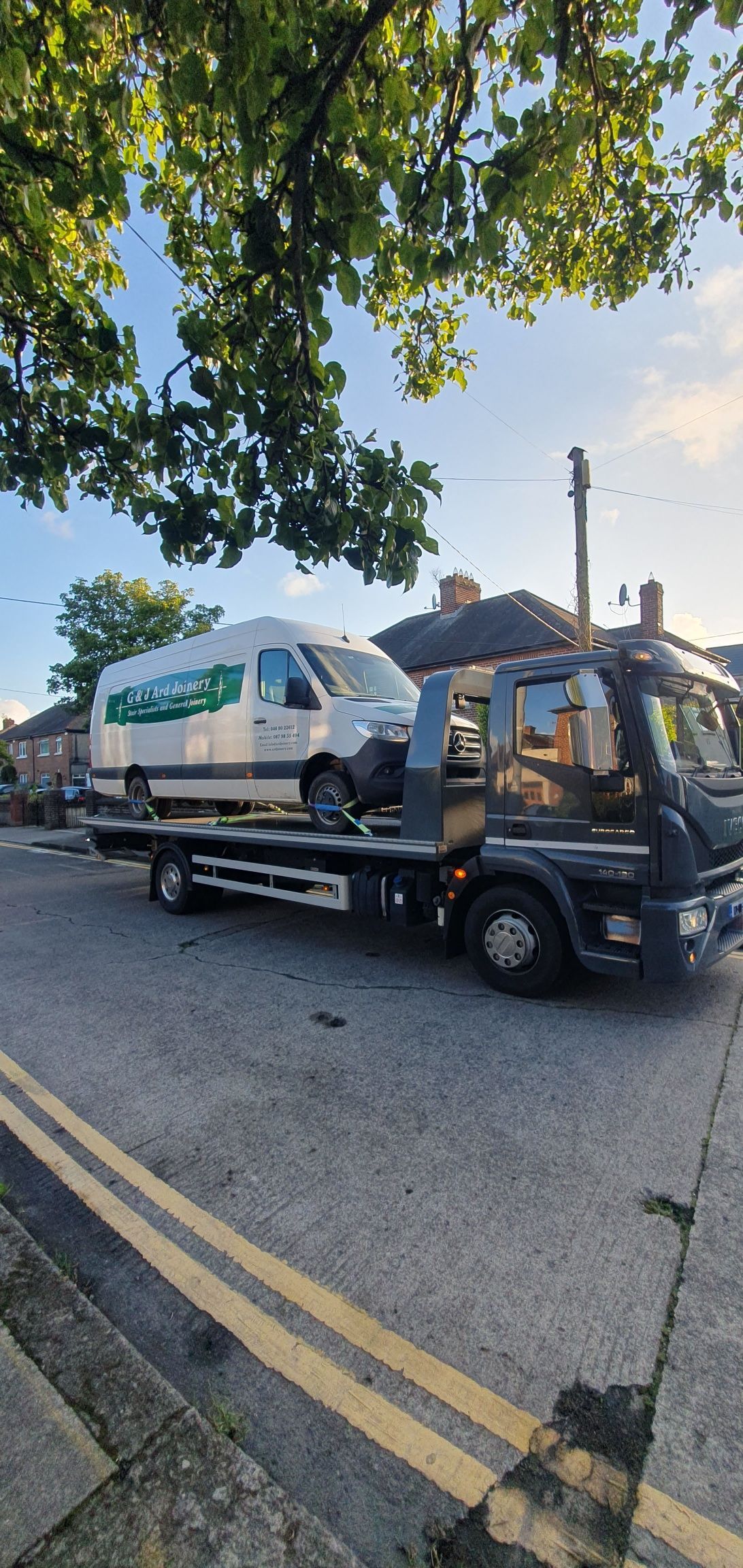 Tow truck carrying a white van parked on a tree-lined street near double yellow lines
