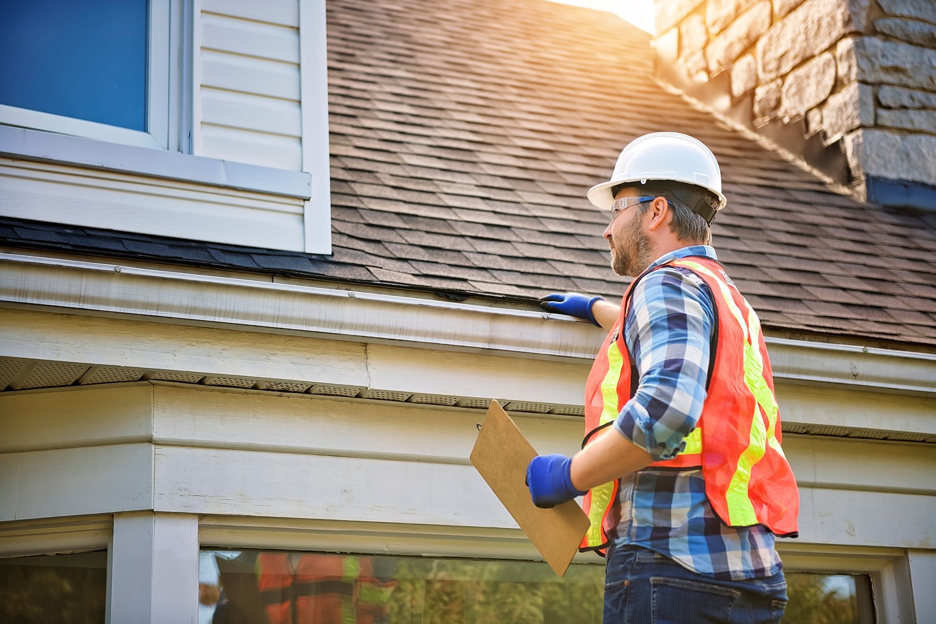 Professional roofing contractor in safety gear inspecting asphalt shingles on a home's roofline. Professional roofing contractor in safety gear inspecting asphalt shingles on a home's roofline.