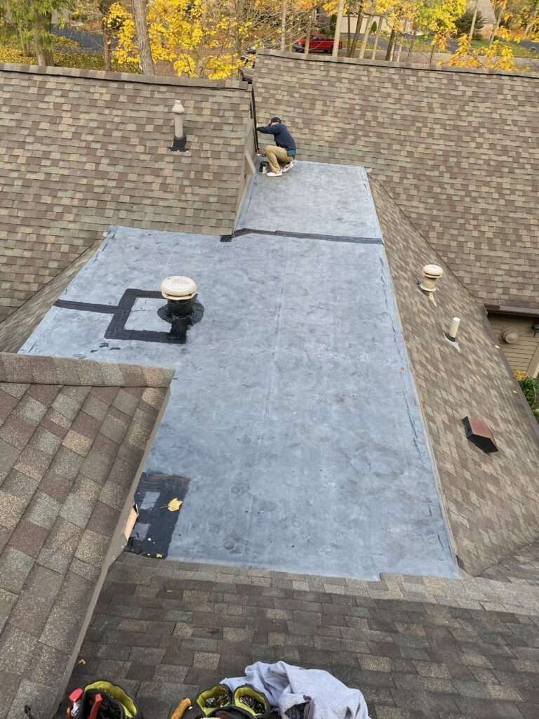 A person on a roof installing flat roofing material; brown shingles surround.