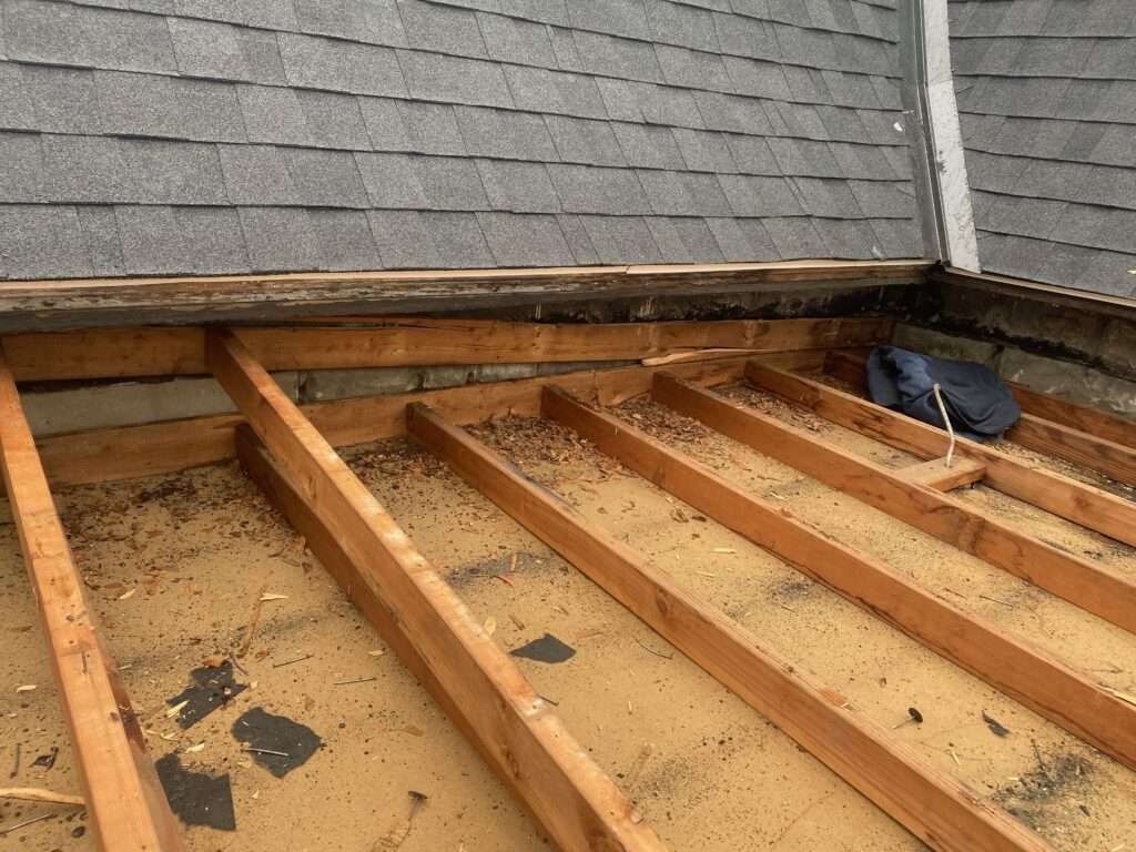 Roof structure exposed, wooden beams, debris, adjacent to shingled roof, angled view.