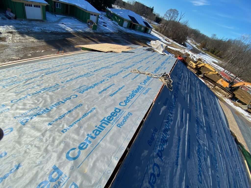 Rooftop covered with CertainTeed Roofing underlayment, houses and construction equipment visible, snow on ground.