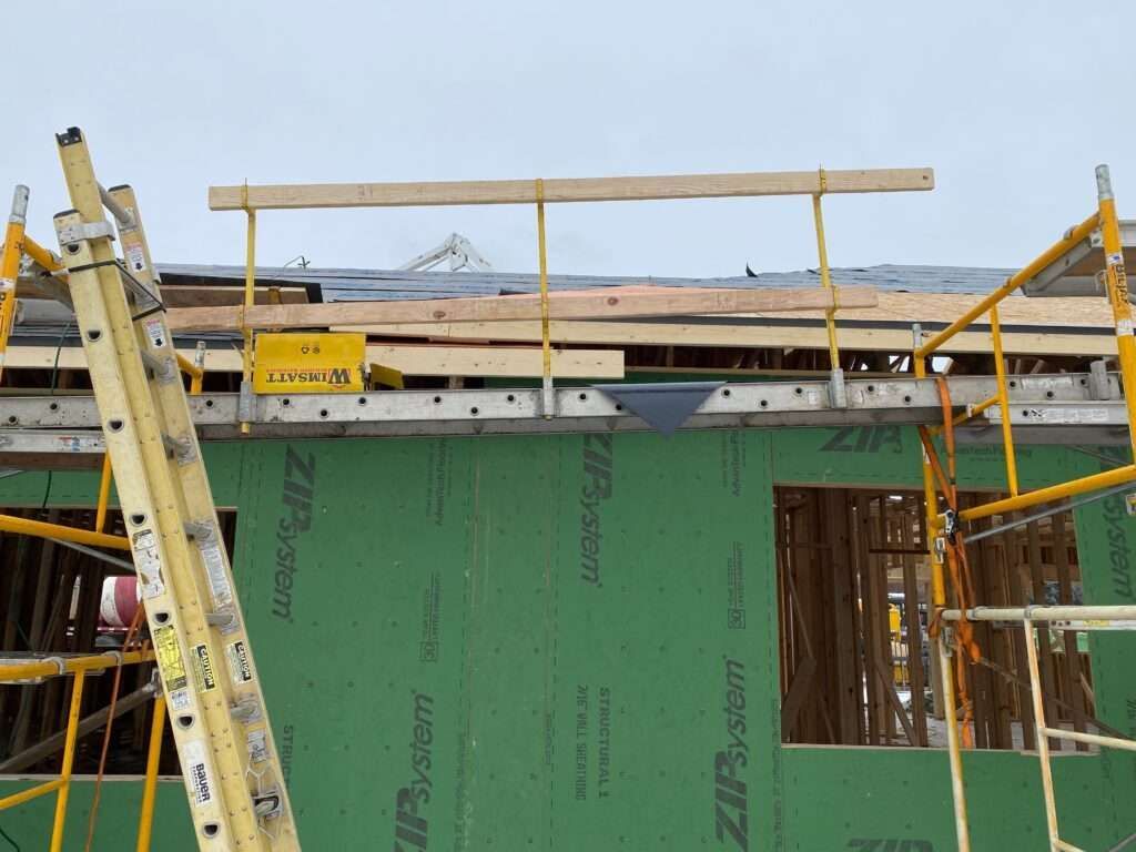 Construction site with scaffolding and safety railings on a roof. Green siding and a ladder are visible.