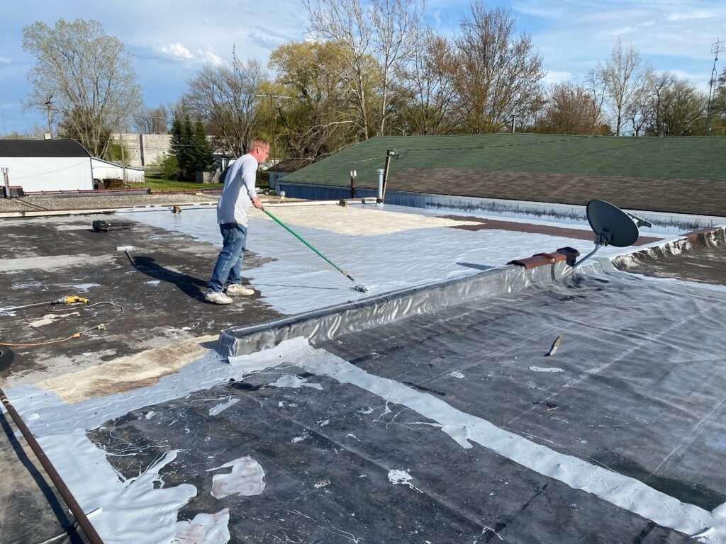 Person applying sealant to a flat roof with a roller. Partially coated roof, clear sky, trees in background.