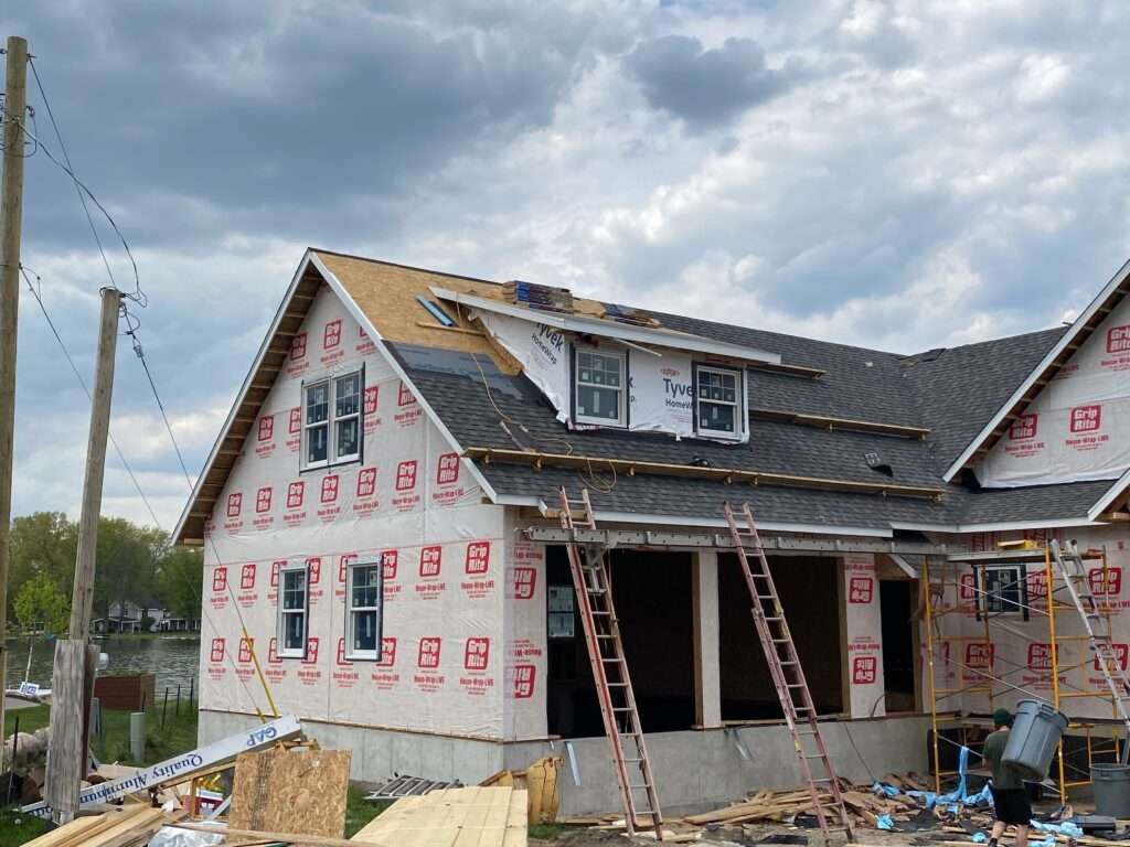 House under construction with exposed sheathing, windows, and ladders, against a cloudy sky.