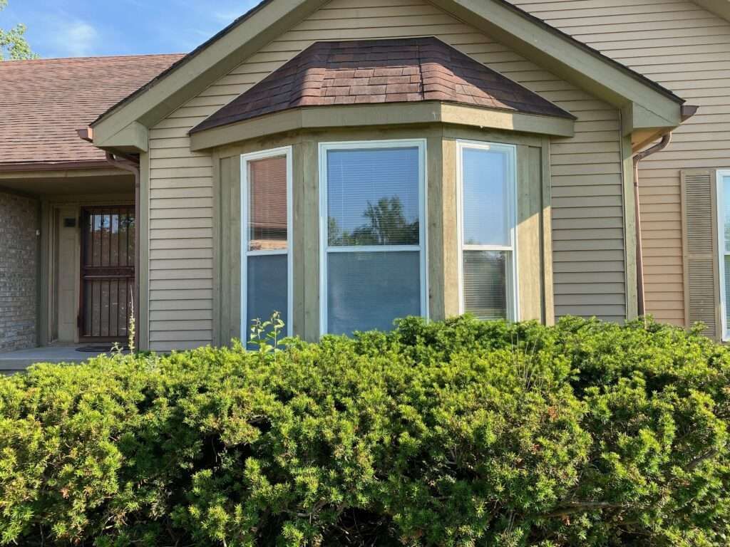 Beige house with a bay window, brown roof, and green bushes.