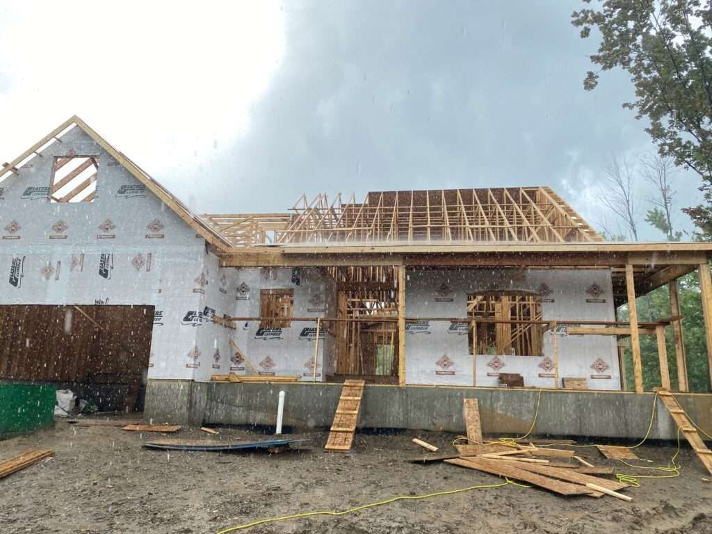 House under construction; wooden frame and roof, covered in wrap, under rainy skies.