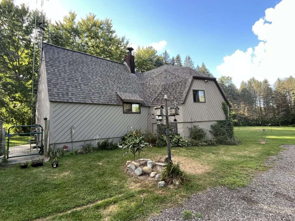 House with gray angled siding, dark shingled roof, and chimney. Lawn in front with bird feeder and trees in background.