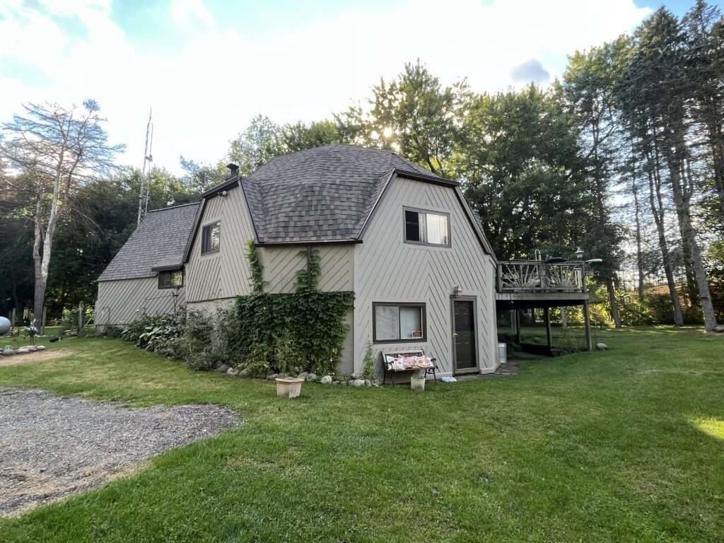 Tan two-story house with a wooden deck and shingled roof, surrounded by green grass and trees.