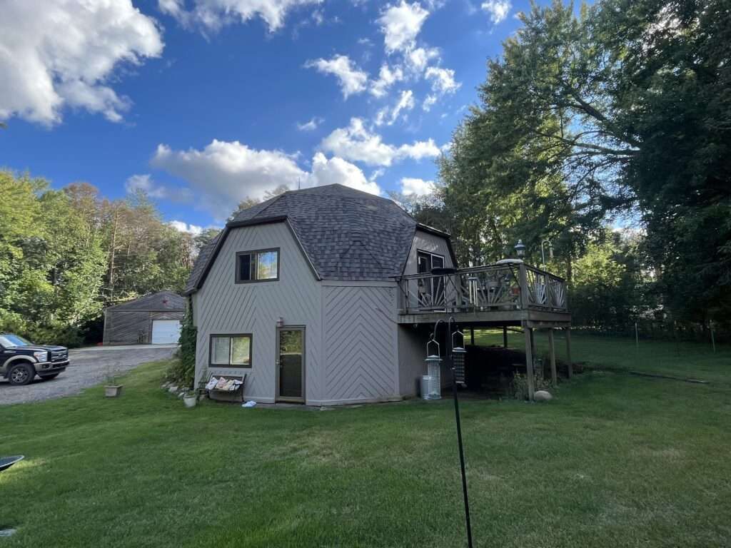 Barn-shaped house with gray siding, dark roof, deck, and green lawn under a partly cloudy sky.