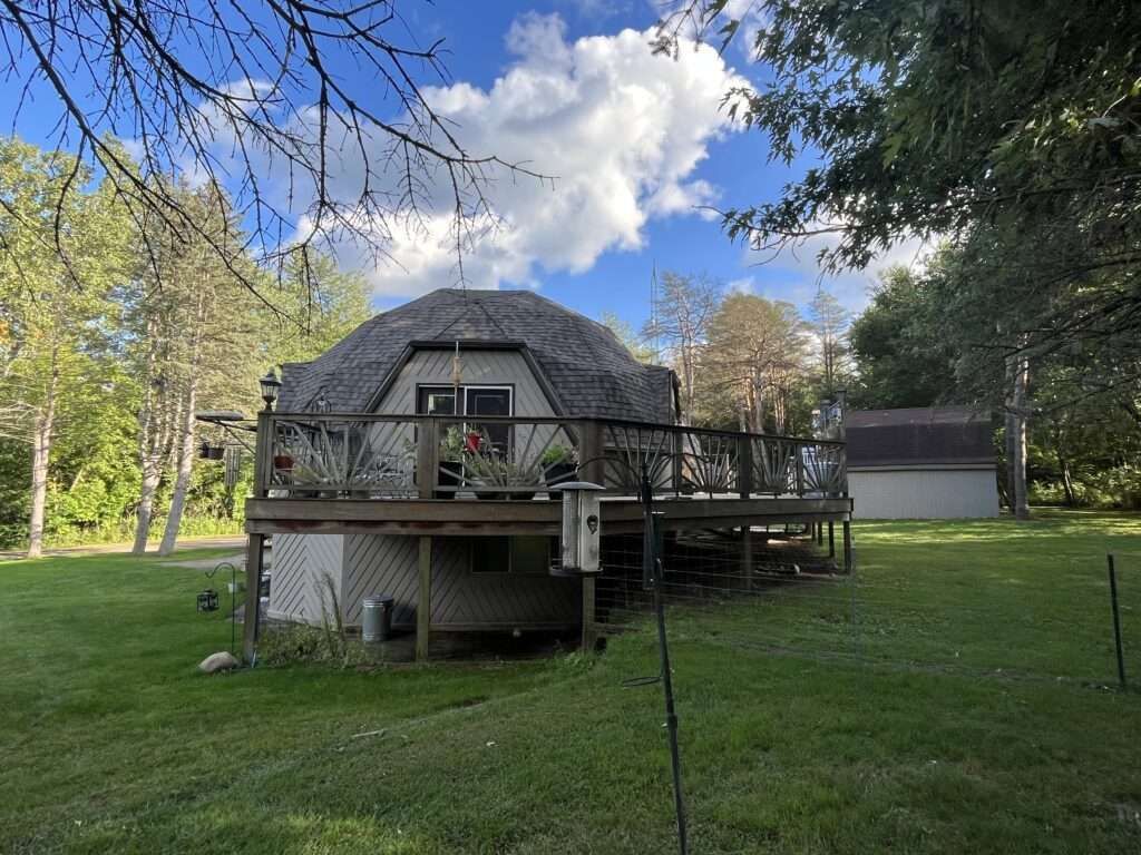 Dome-shaped house with a large deck, surrounded by trees and a green lawn under a partly cloudy sky.