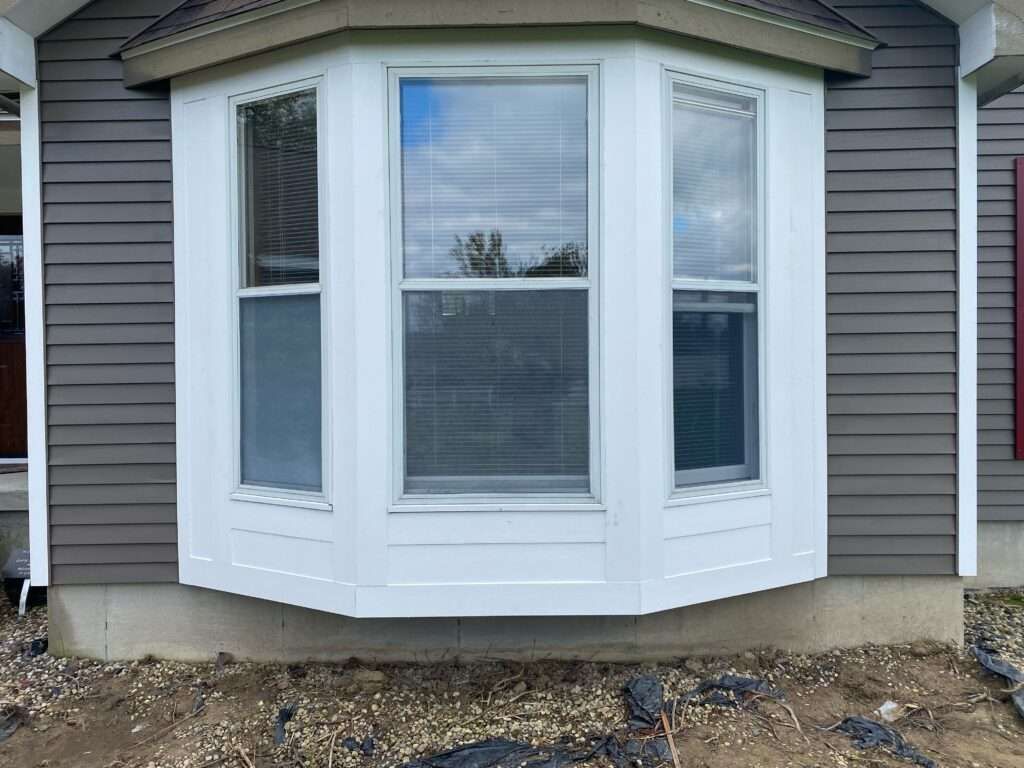 Bay window on a house with white trim and gray siding; dirt and landscaping in front.