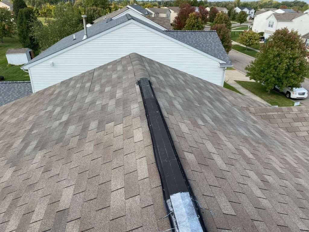 Overhead view of a roof with brown shingles, a black vent, and surrounding houses in a residential area.