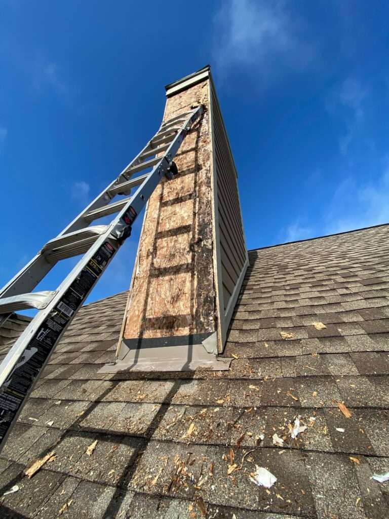 Ladder leans against a chimney with exposed wood siding on a shingled roof, blue sky above.