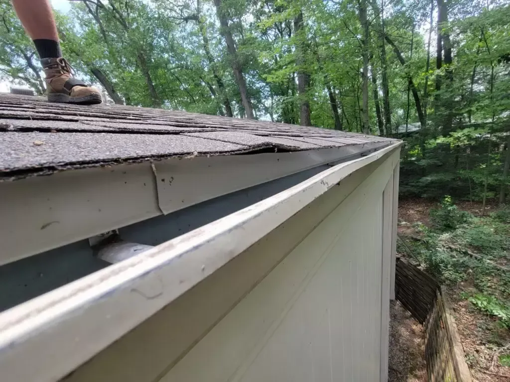 Person on roof next to a damaged gutter, with trees in the background.