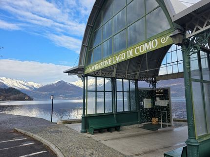 Una stazione degli autobus verde con un lago e montagne innevate sullo sfondo, visibile la scritta "LAGO DI COMO".