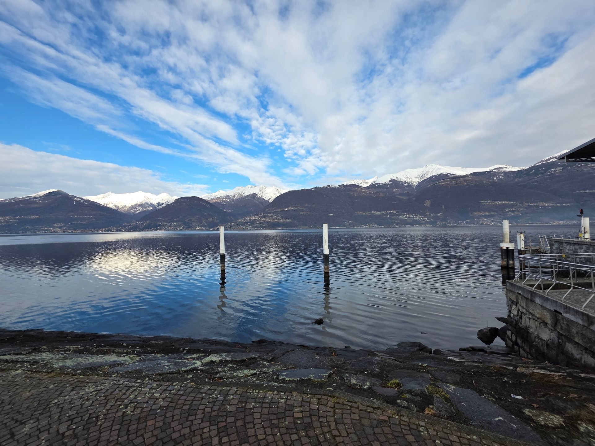 Paesaggio lacustre con montagne, acqua riflettente, cielo azzurro e nuvole bianche. Un molo in primo piano a destra.