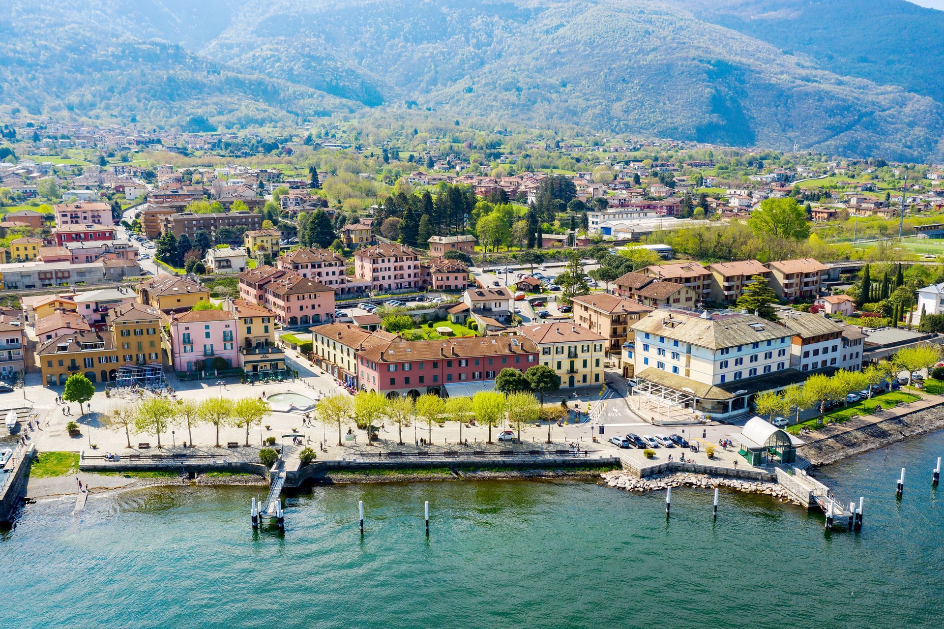 Città sul lago con edifici colorati, un lungomare alberato e montagne sullo sfondo.