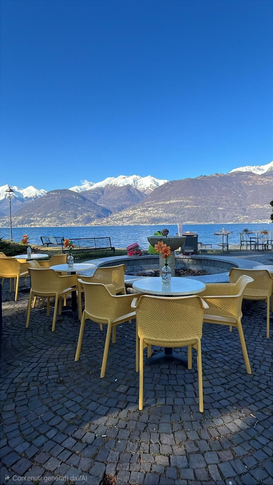 Bar all'aperto con sedie gialle, tavoli rotondi, lago e montagne innevate sotto un cielo azzurro.