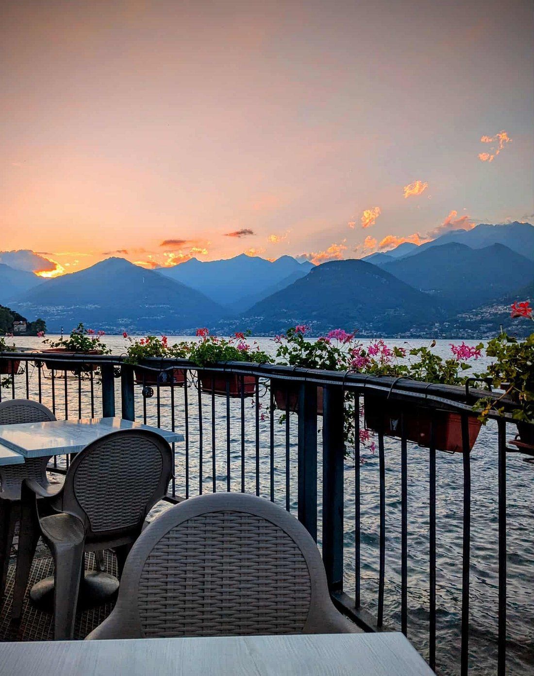 Tramonto sul lago e sulle montagne, visto dal patio del ristorante. Sedie, tavoli, fioriere e una ringhiera sono in primo piano.