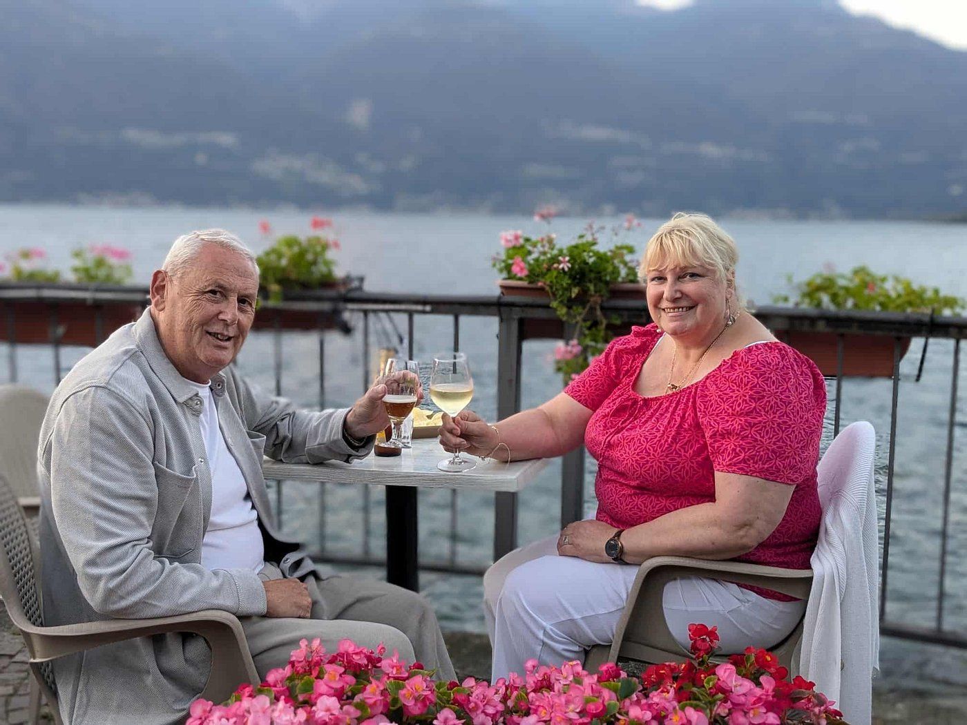 Coppia che brinda in un bar sul lago. Uomo con giacca leggera, donna con top rosa, fiori in primo piano.