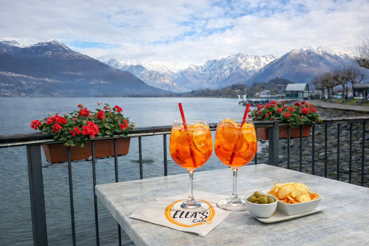 Due Aperol Spritz su un tavolo in riva a un lago con le montagne sullo sfondo. Si vedono anche olive e patatine fritte.