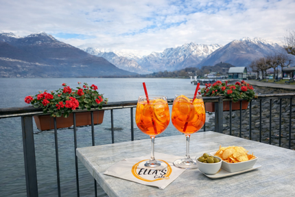 Due Aperol Spritz su un tavolo con un lago e montagne sullo sfondo. Si vedono anche olive e patatine fritte.