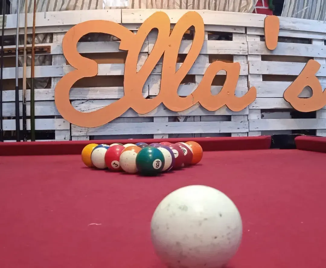 Palle da biliardo disposte su un tavolo di feltro rosso, con la palla bianca in primo piano e il cartello "Ella's" sullo sfondo.